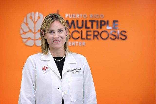 A white, blonde woman smiles, standing in front of a wall that says "Puerto Rico Multiple Sclerosis Center."