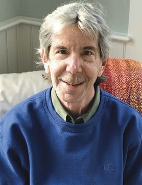 A person with gray hair and a mustache sits on a couch and smiles