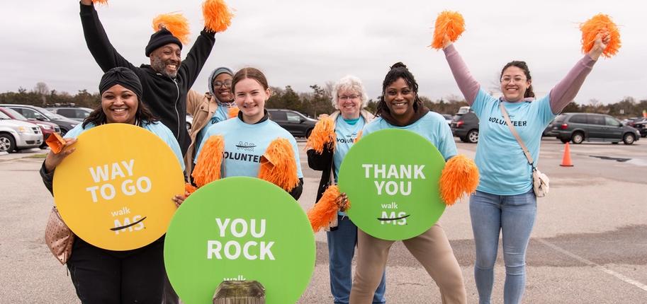 A group of Walk MS 2024 volunteers in blue t-shirts holding orange pom-poms and three circular signs reading "Way to Go," "You Rock" and "Thank You"
