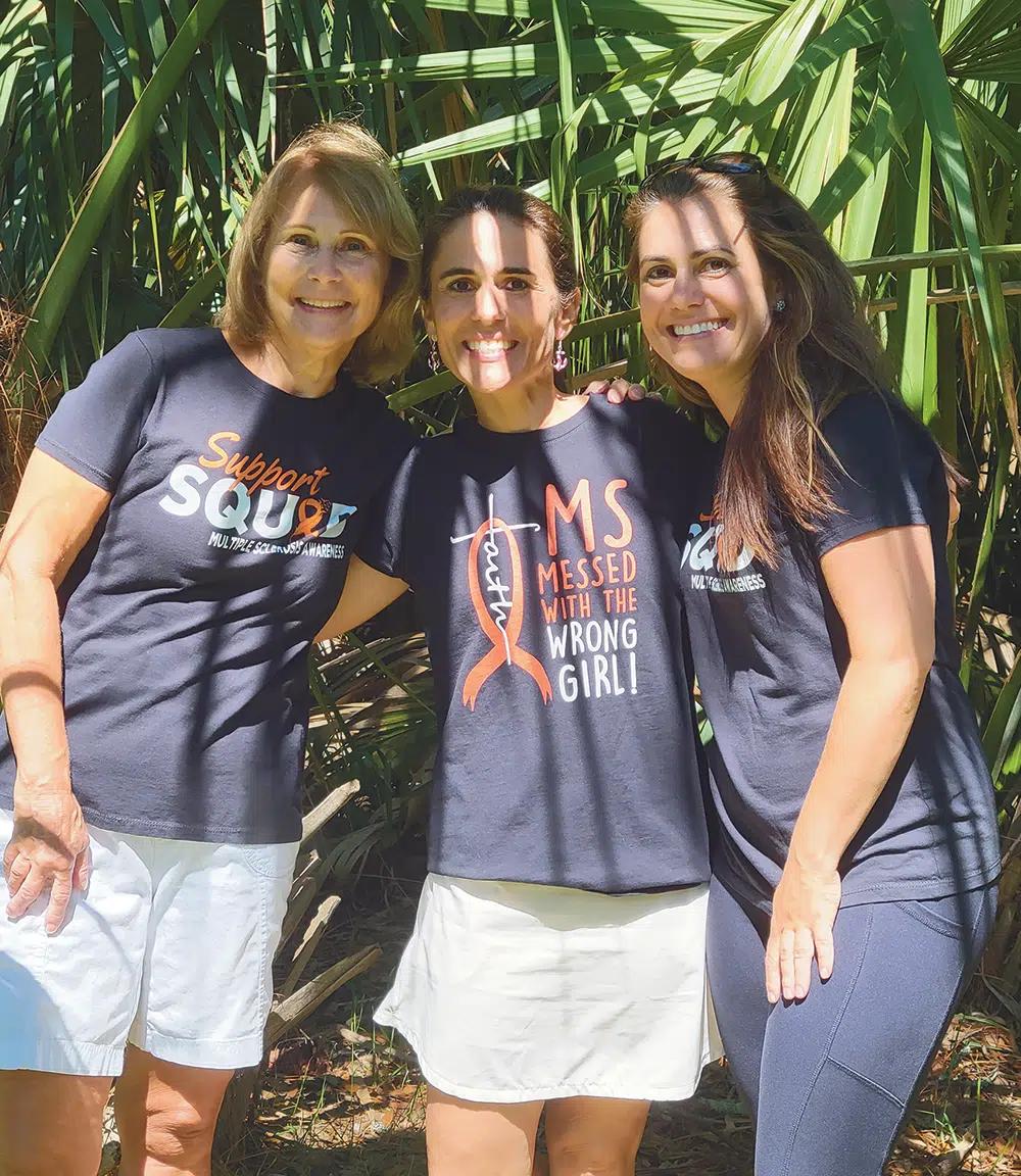 Shelly Krebs stands between two women. All three have their arms around each other and wear MS awareness shirts.