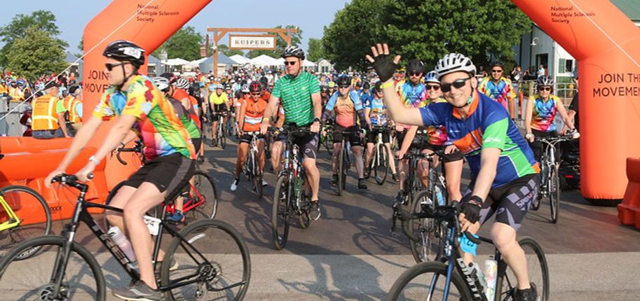 A large group of cyclists pass under an orange arch at the finish line of Bike MS.