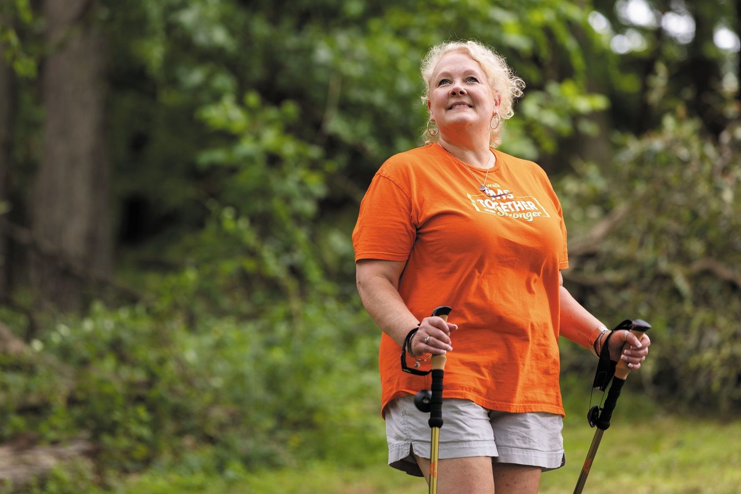 Samantha Opferman wears an orange shirt and holds hiking poles while standing outside and smiling.