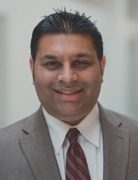 A brown-skinned man with black hair wearing a brown suit, white shirt, and red and brown tie.