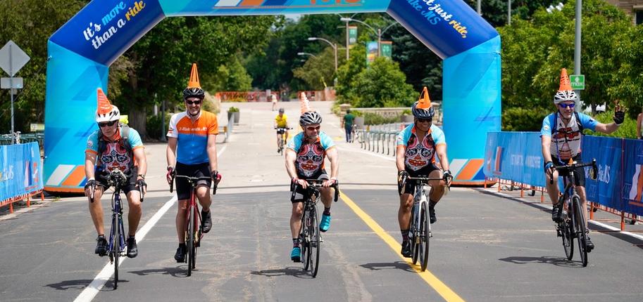 Five cyclists ride side by side with the blue Bike MS starting arch behind them.