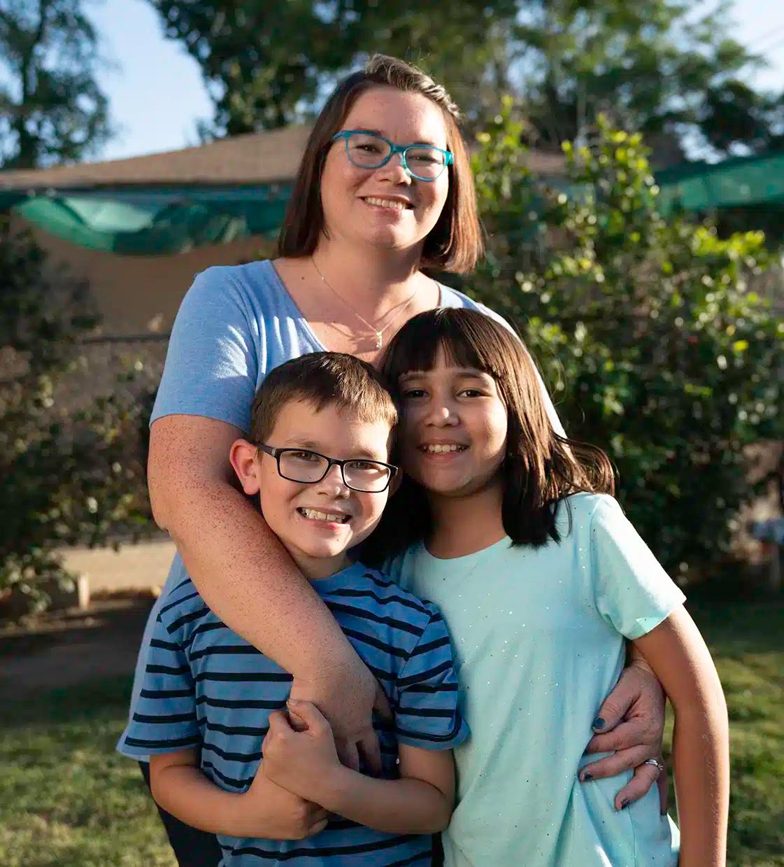 Sarah Quezada stands with her arms around her two kids. Her son stands on the left wearing a blue striped shirt and glasses while her daughter stands to the right wearing a light blue shirt with sparkles.