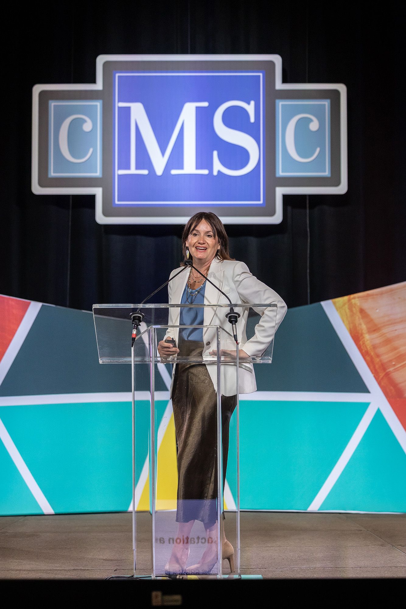A woman in business attire speaks on a podium in front of a colorful background that reads CMSC.