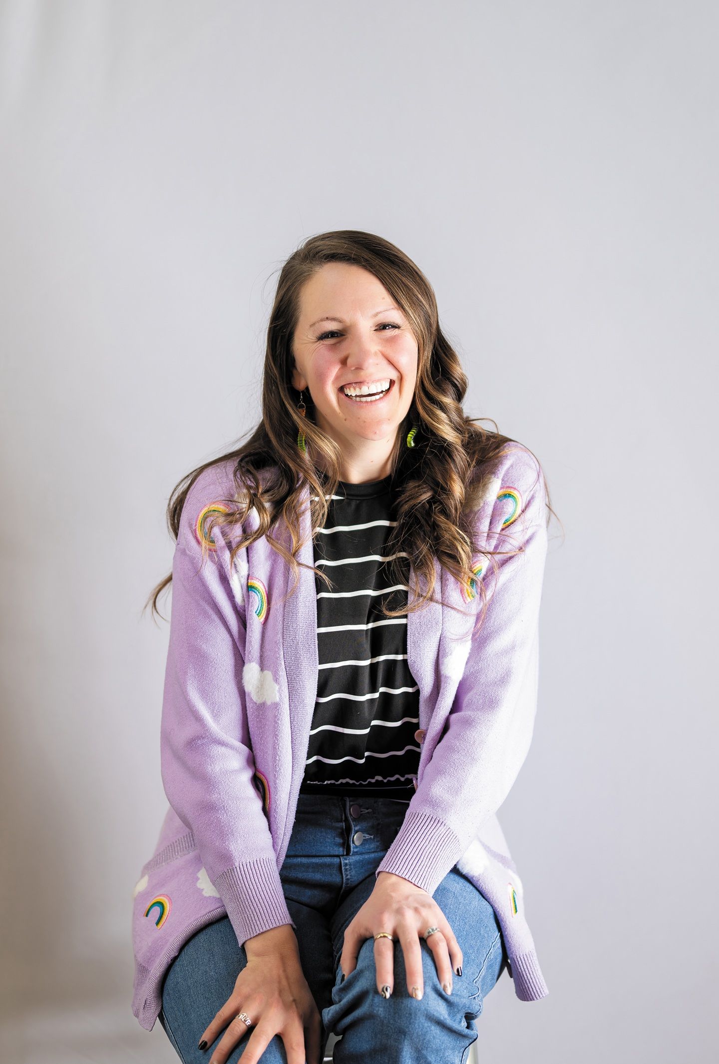 A white woman with long, brown hair sits smiling in jeans, a pink jacket and black and white shirt.