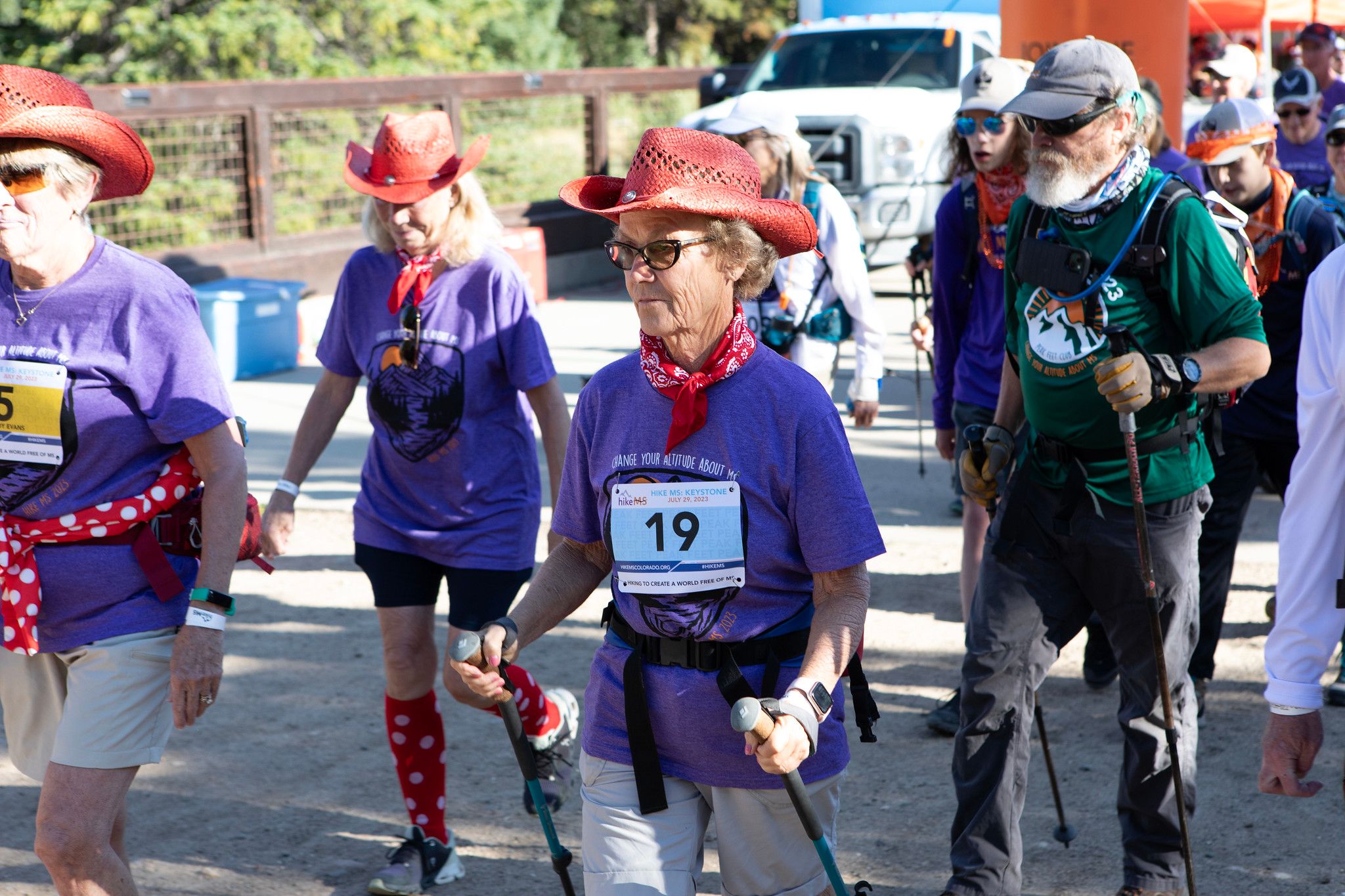 A woman in a purple shirt holding hiking poles walks a trail during Hike MS surrounded by other participants.