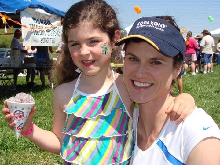 Photo of a young daughter with a butterfly painted on her face, holding a snowcone, wraps her arm around her smiling mother, wearing a visor, at a Walk MS: Nashville event.