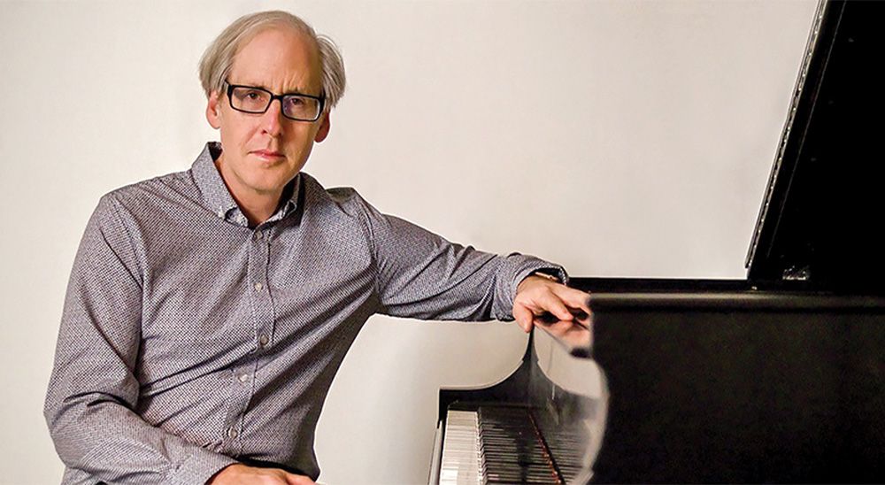 An older, white man with glasses sits at a grand piano.