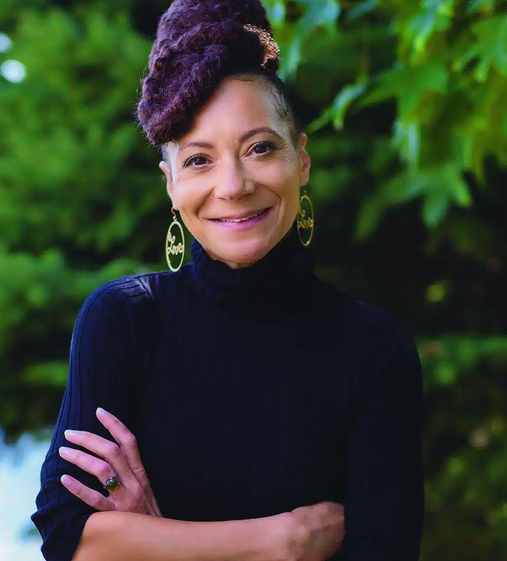 A woman stands with her arms folded across her front, smiling at the camera. She wears a black turtleneck and gold earrings, with her hair styled up.
