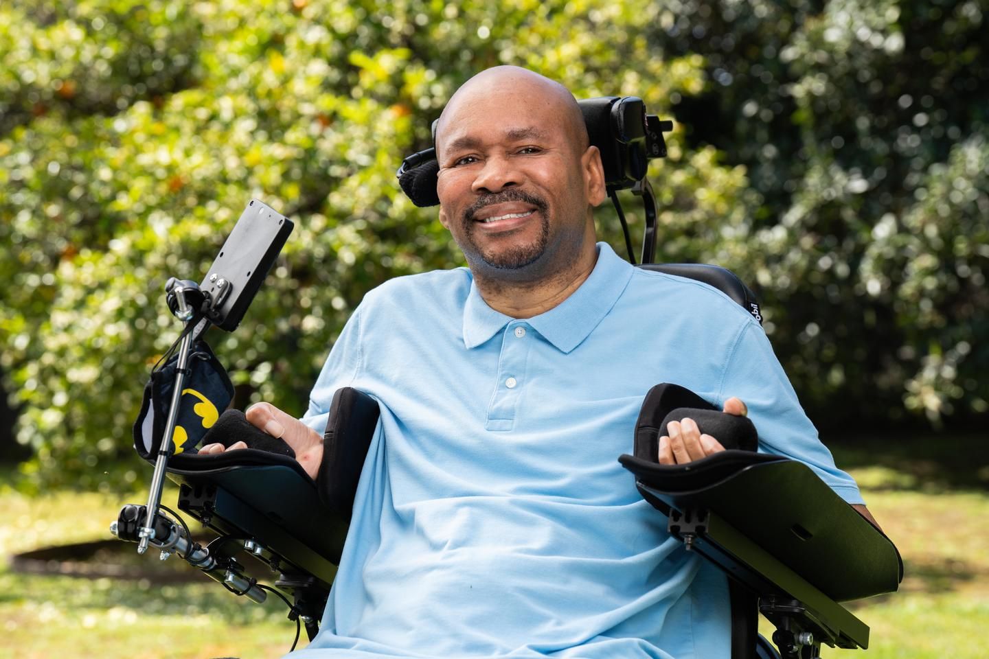 A man who lives with MS sits in his wheelchair in this backyard, smiling.