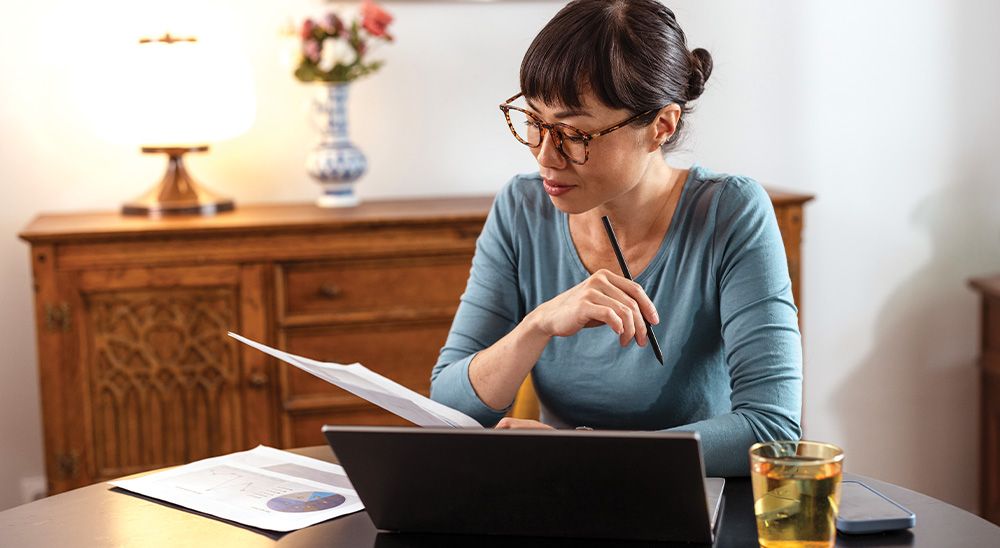 A woman sits at a desk, looking at a laptop and paperwork.