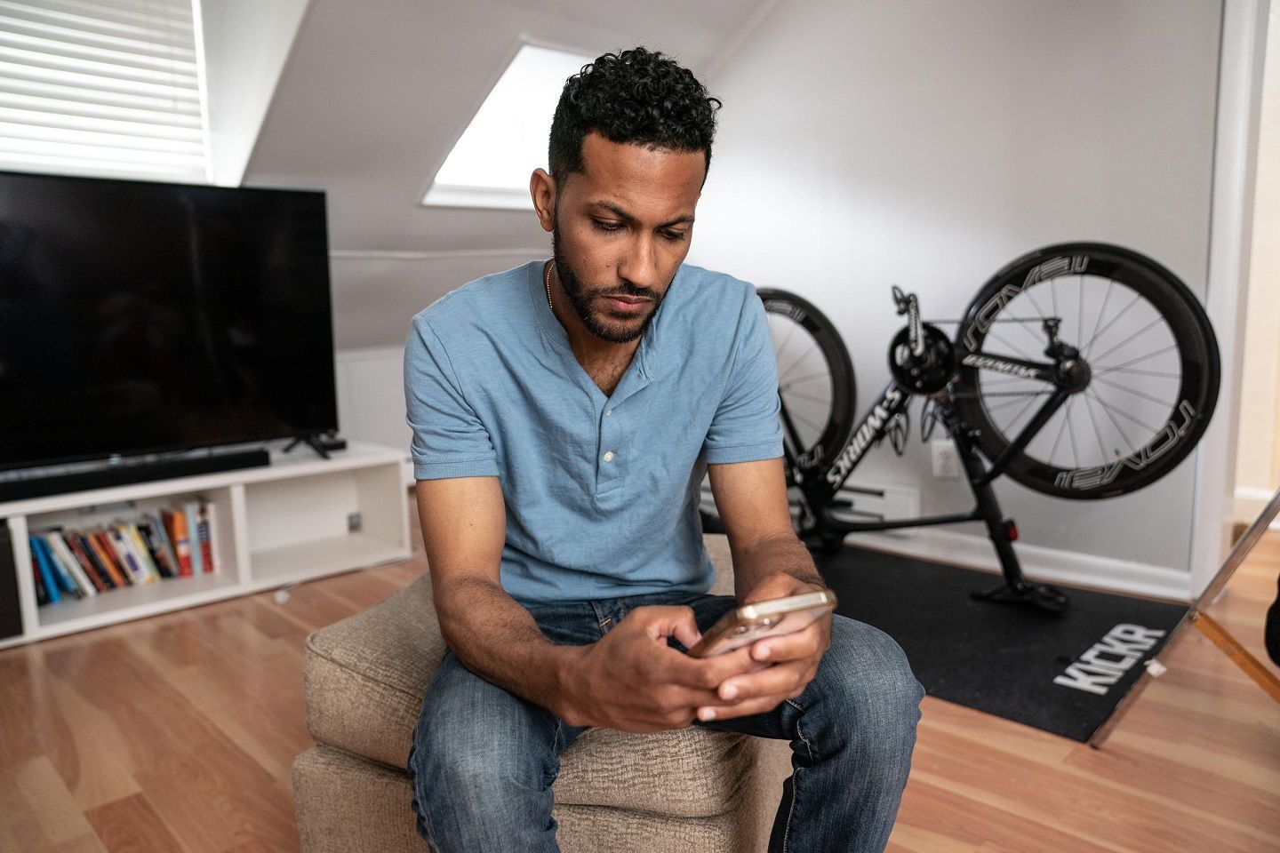 A man sitting down in front of a TV and up-side-down bicycle looks at his cell phone.
