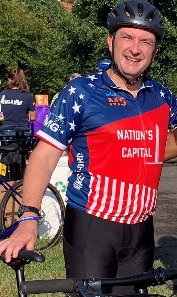A man in a cycling helmet and a red, blue and white jersey that reads Nation’s Capital smiles while holding his bike outdoors.