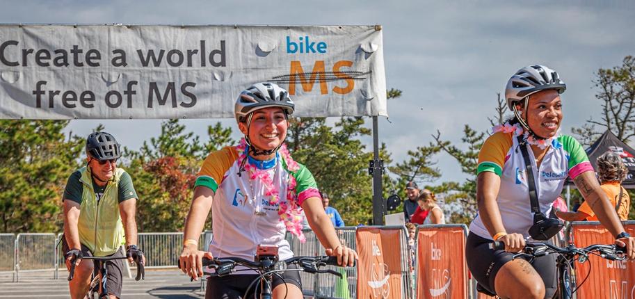 Three cyclists ride their bikes along a road with a sign that reads "Create a world free of MS - bike MS" behind them.