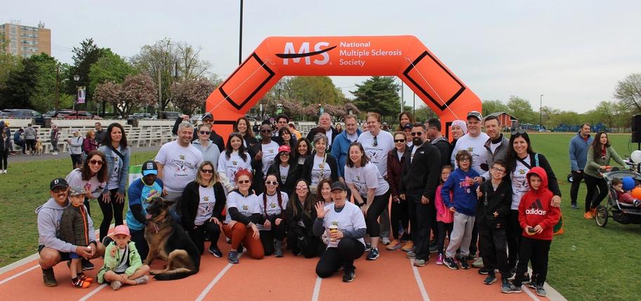 A large group of people pose smiling on a running track in front of an orange Walk MS arch.