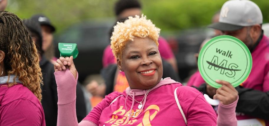 A black woman with short blonde hair and wearing a pink shirt, smiles towards the camera while holding a green Walk MS sign.