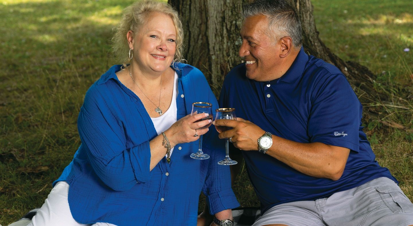 A white woman and her husband sit outside under a tree, touching glasses of wine in a âcheersâ motion.