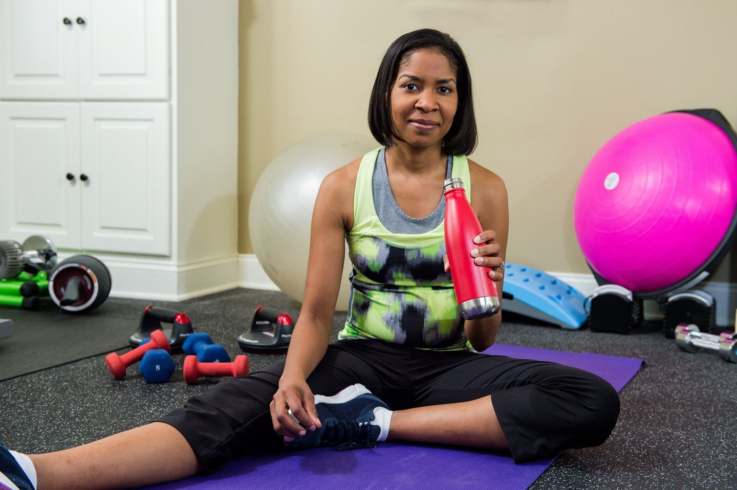 A woman in exercise clothes sits drinking water in a workout room.