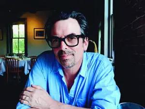 White man with dark hair and short goatee and black glasses.