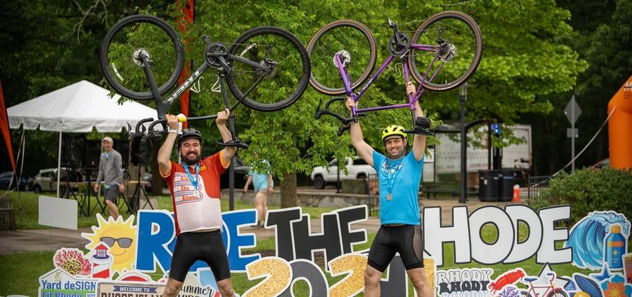 Two cyclists stand in front of a sign that reads "Ride the Rhode 2024" with their bikes raised above their heads.