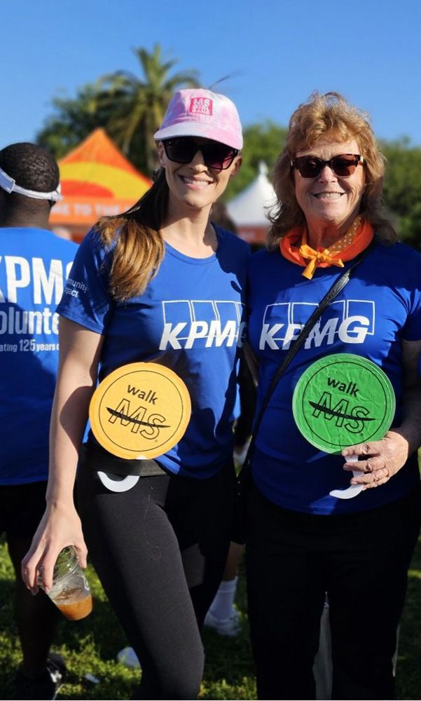 Two women in blue T-shirts that read KPMG pose for a picture holding yellow and green Walk MS circles during an outdoor event.