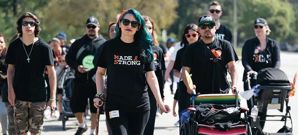 A young woman with blue hair, wearing a "Made Strong" shirt and using a cane walks in the front of a crowd of people.