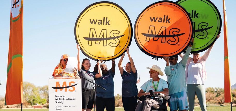 A group of people stand on stage at Walk MS with three large yellow, orange and green circular signs that read "Walk MS" raised above their heads.