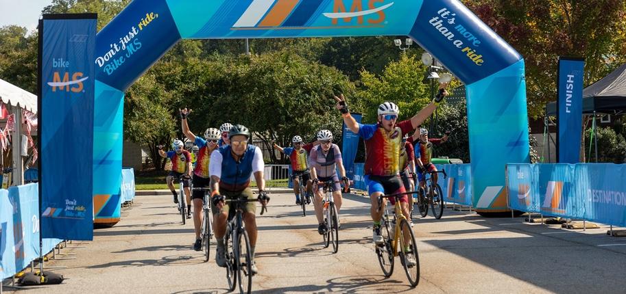 A group of cyclists in two lines pass under a blue Bike MS finish line arch.