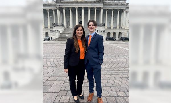 A white woman in a black suit and orange shirt stands in front of a government building beside a white man wearing a blue suit and orange tie.