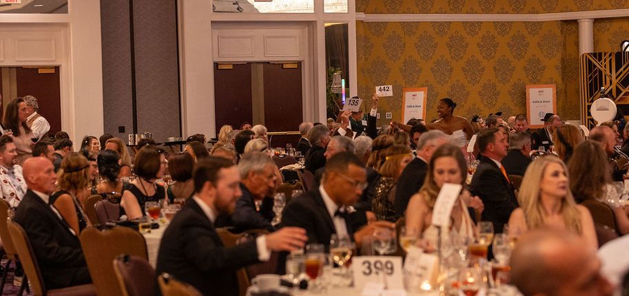 A ballroom of people in evening wear sitting at tables with drinks.