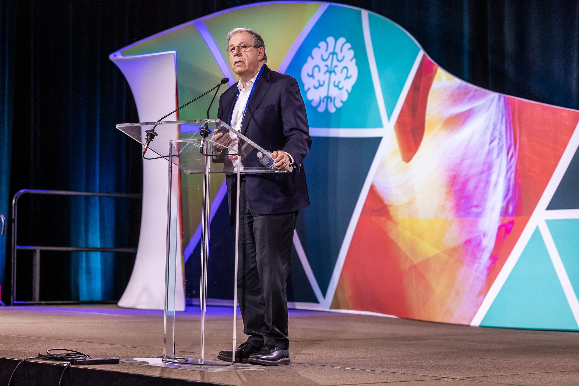 A man stands at a clear podium on stage, in front of a colorful background.