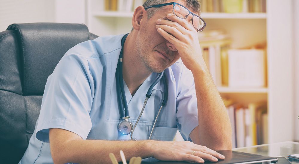 A doctor sits at a desk with his hand covering his face.