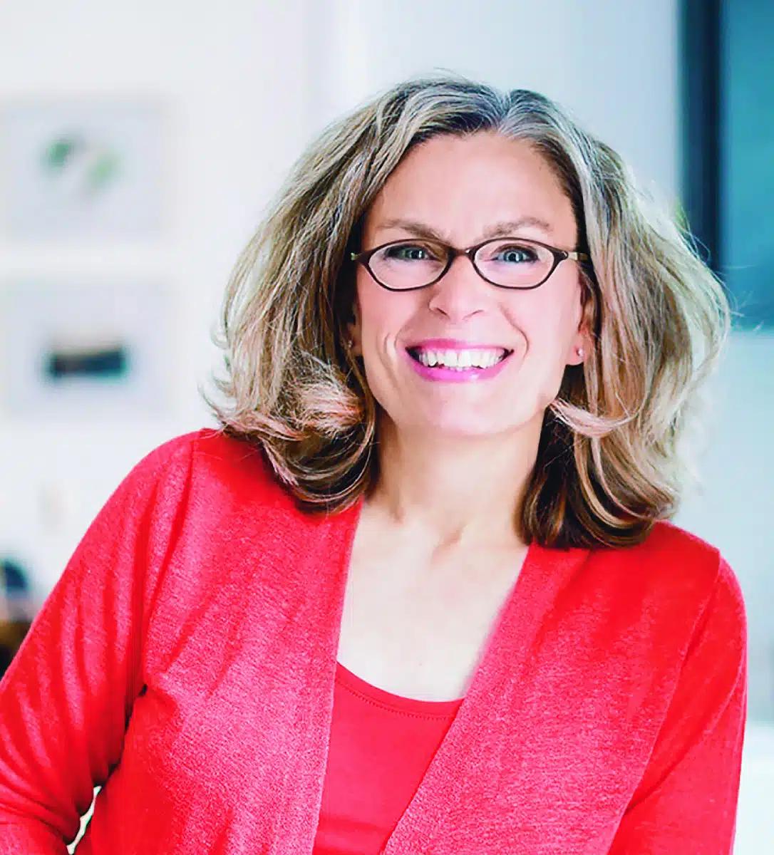 A Caucasian woman with shoulder-length hair and glasses smiles at the camera