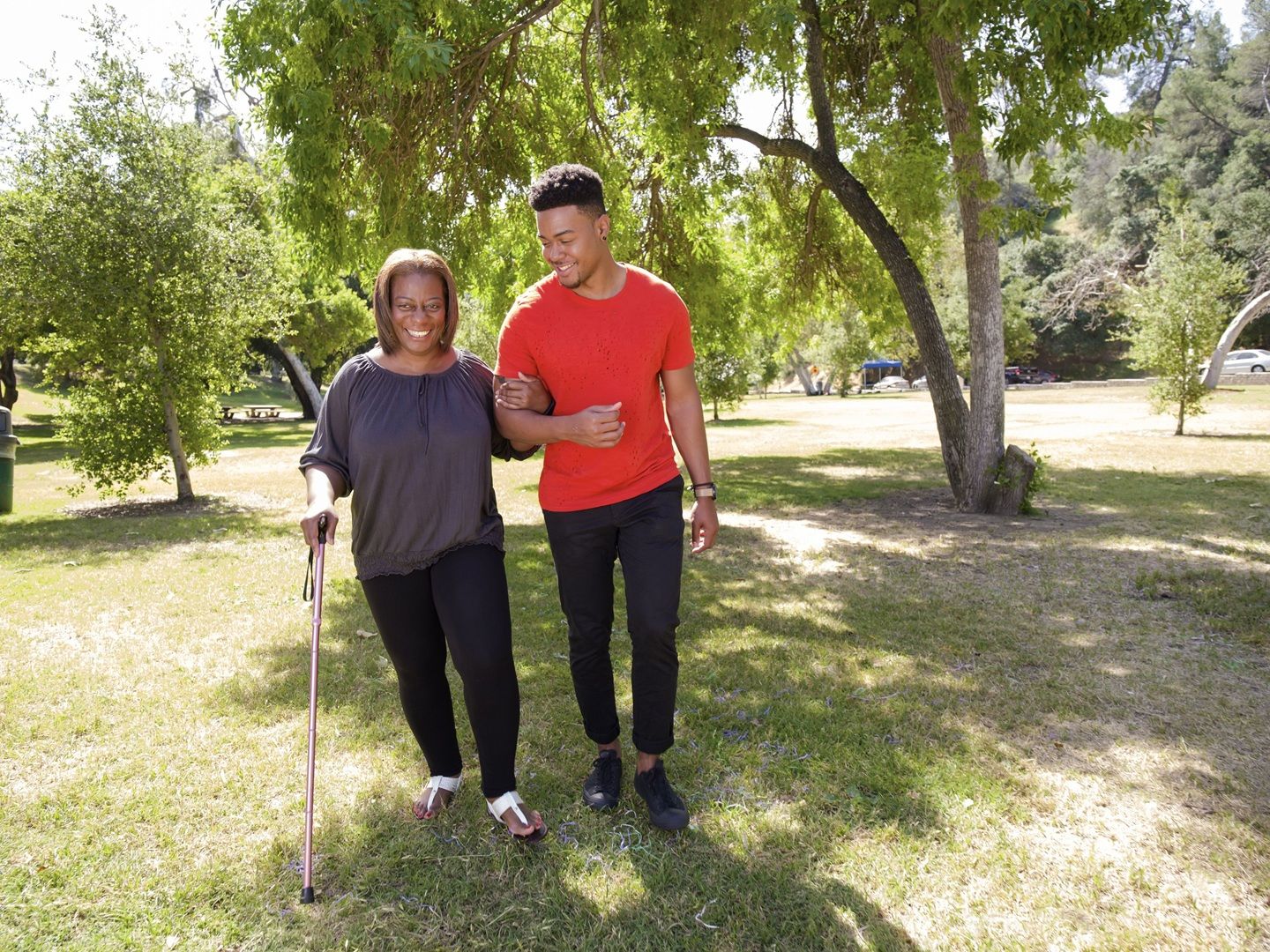 A dark-skinned person with shoulder-length brown hair using a cane walks outside arm-in-arm with a tall, dark-skinned person with short brown hair.