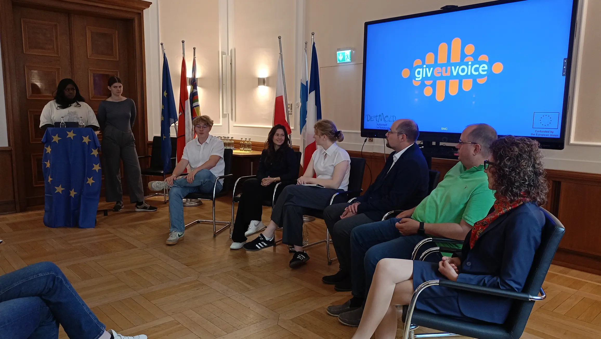 Debata z politykami w sali ratusza w Detmoldzie, zorganizowana w ramach projektu European Youth Campus. Na zdjęciu widać uczestników siedzących w rzędzie krzeseł, słuchających prelegenta. Z tyłu za panelem dyskusyjnym znajduje się ekran z logo projektu „give eu voice” oraz szereg flag, w tym Unii Europejskiej i Niemiec. Po lewej stronie, na mównicy ozdobionej flagą UE, stoi osoba prowadząca wydarzenie. W tle widać zabytkowe, drewniane drzwi.