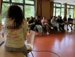 Uczestnicy warsztatów w ramach European Youth Campus w Detmoldzie. Widok z tyłu na osobę w koszulce z napisem „give eu voice” i „TEAM POLAND”, siedzącą na biurku. Naprzeciwko niej, w jasnej sali z dużymi oknami, siedzi w półokręgu grupa młodych ludzi, przysłuchując się prelegentowi. Podłoga jest pomarańczowa.