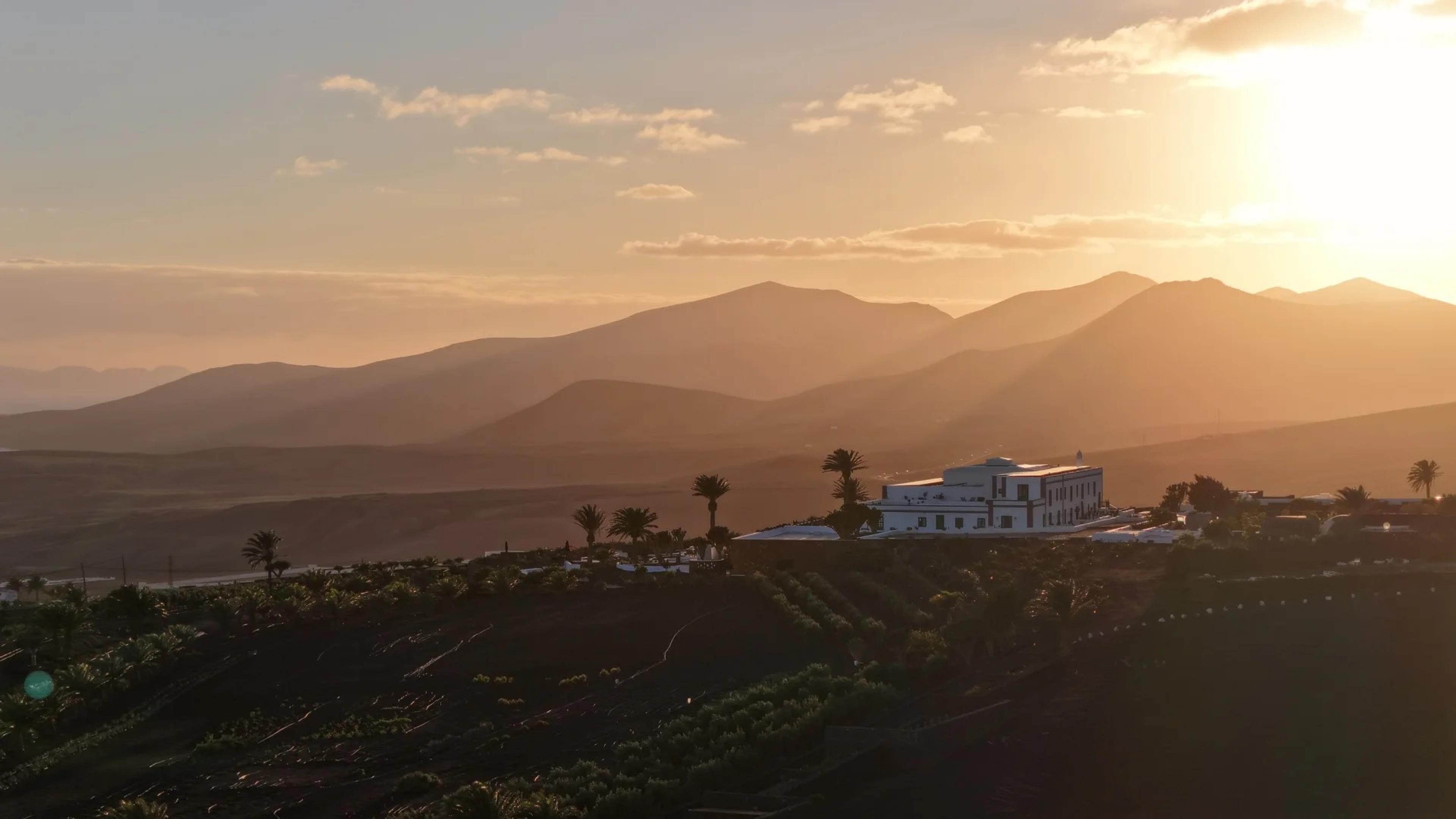 A hillside town in Lanzarote seen from a distance at sunset, with a white building partly visible among palm trees below layered mountains lit orange by the setting sun.