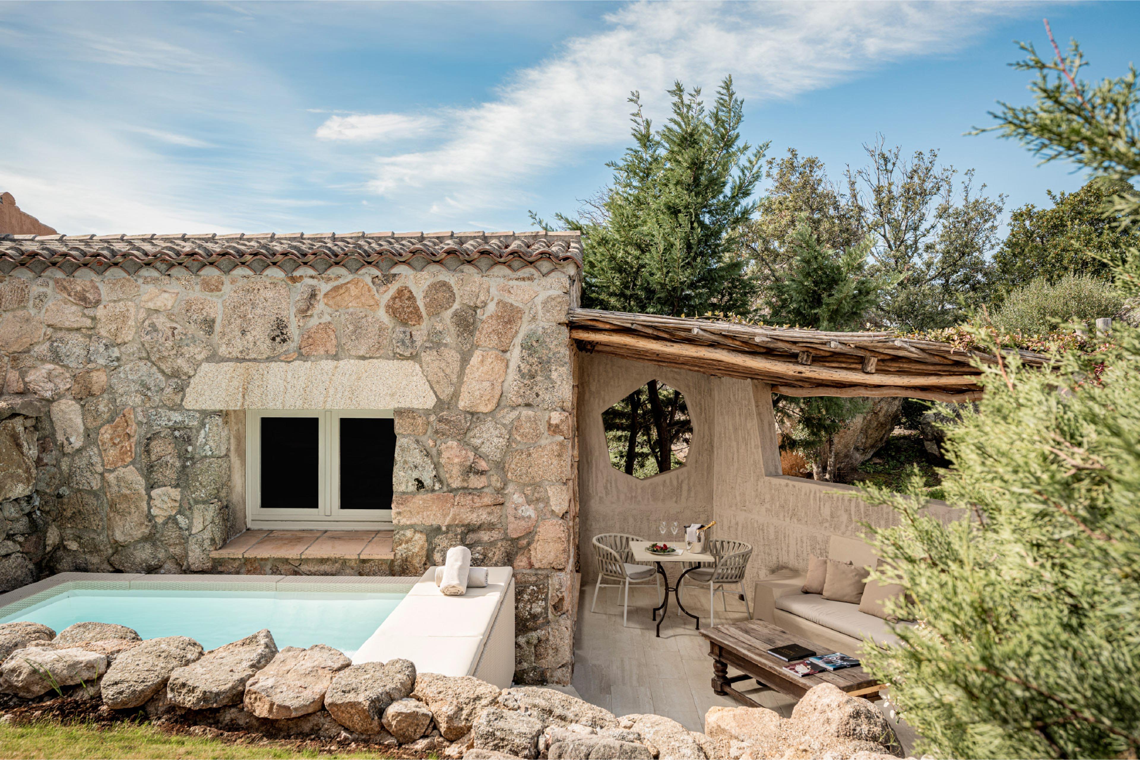 A stone cottage with a tiled roof and shuttered windows, a plunge pool at its base and a covered terrace with a table and sofa to one side.