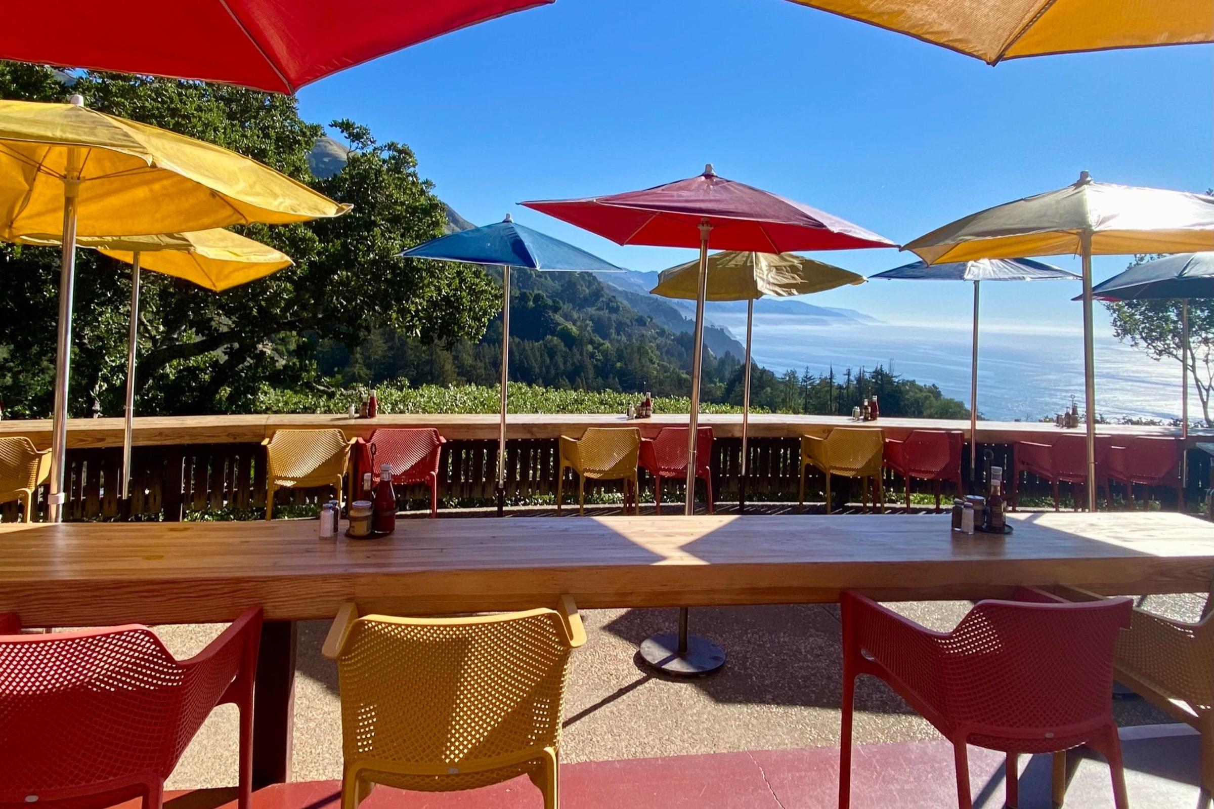 A terrace of tables under red and yellow umbrellas on a cliff, the coast and mountains running away behind.