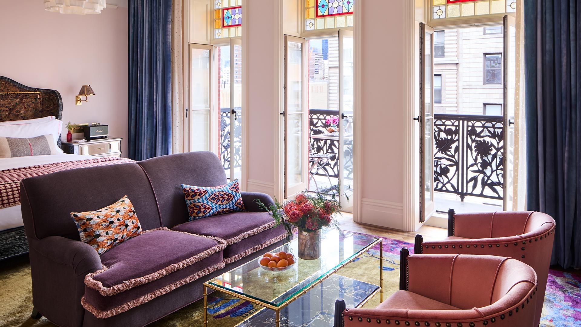 A hotel suite with a purple sofa and armchairs around a glass coffee table, facing tall stained glass doors open to a wrought iron balcony.