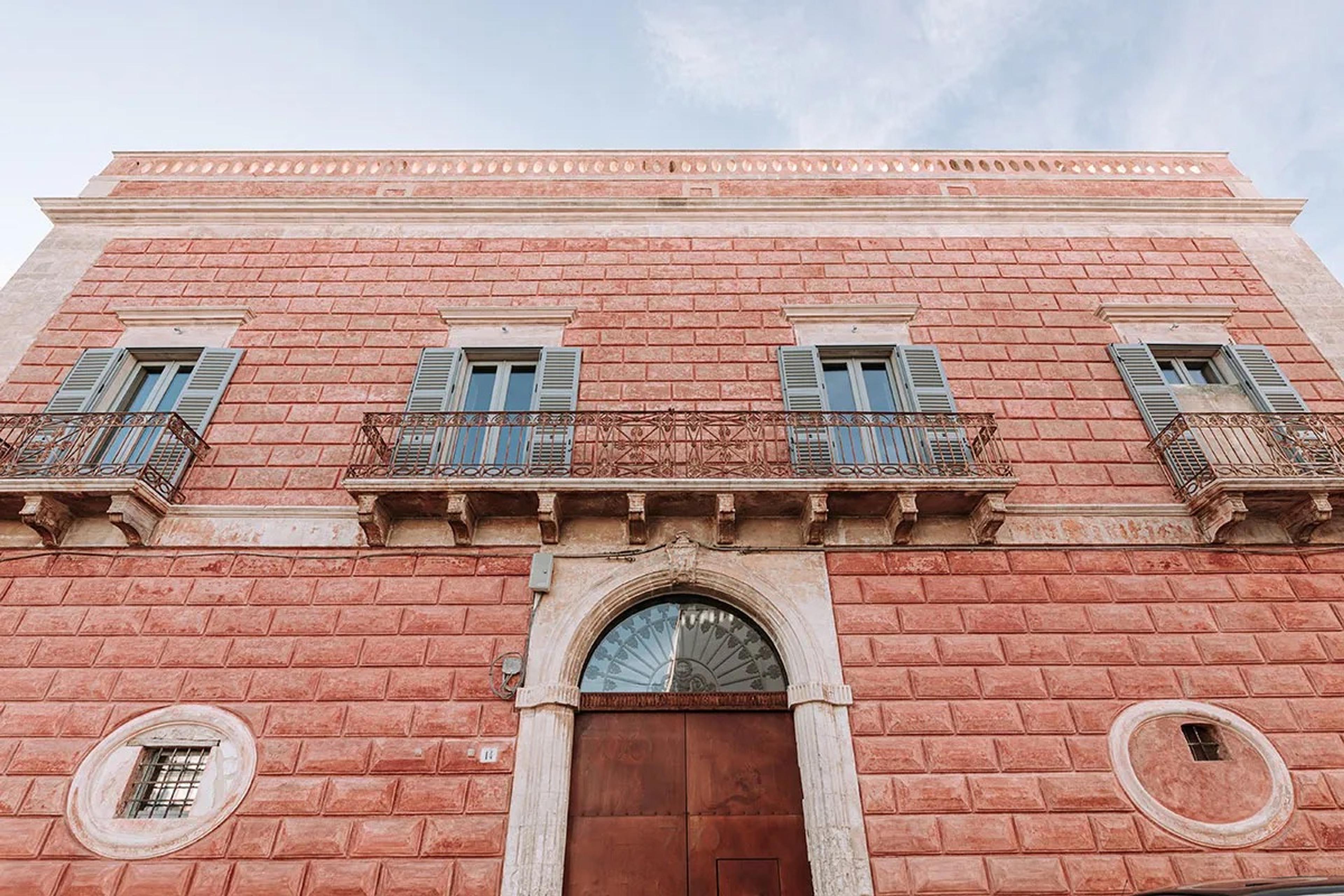 A low-angle view of the hotel's terracotta brick facade, with grey shuttered windows, iron balconies, and an arched stone doorway against a blue sky.