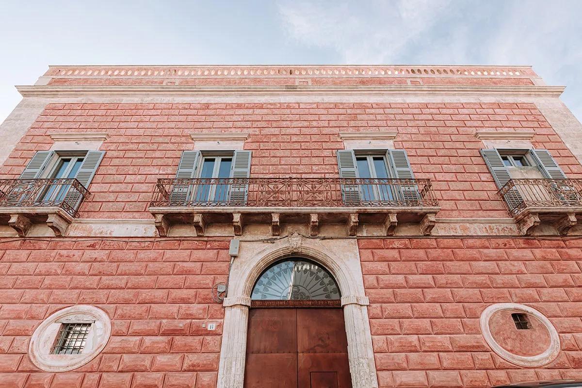 A low-angle view of the hotel's terracotta brick facade, with grey shuttered windows, iron balconies, and an arched stone doorway against a blue sky.