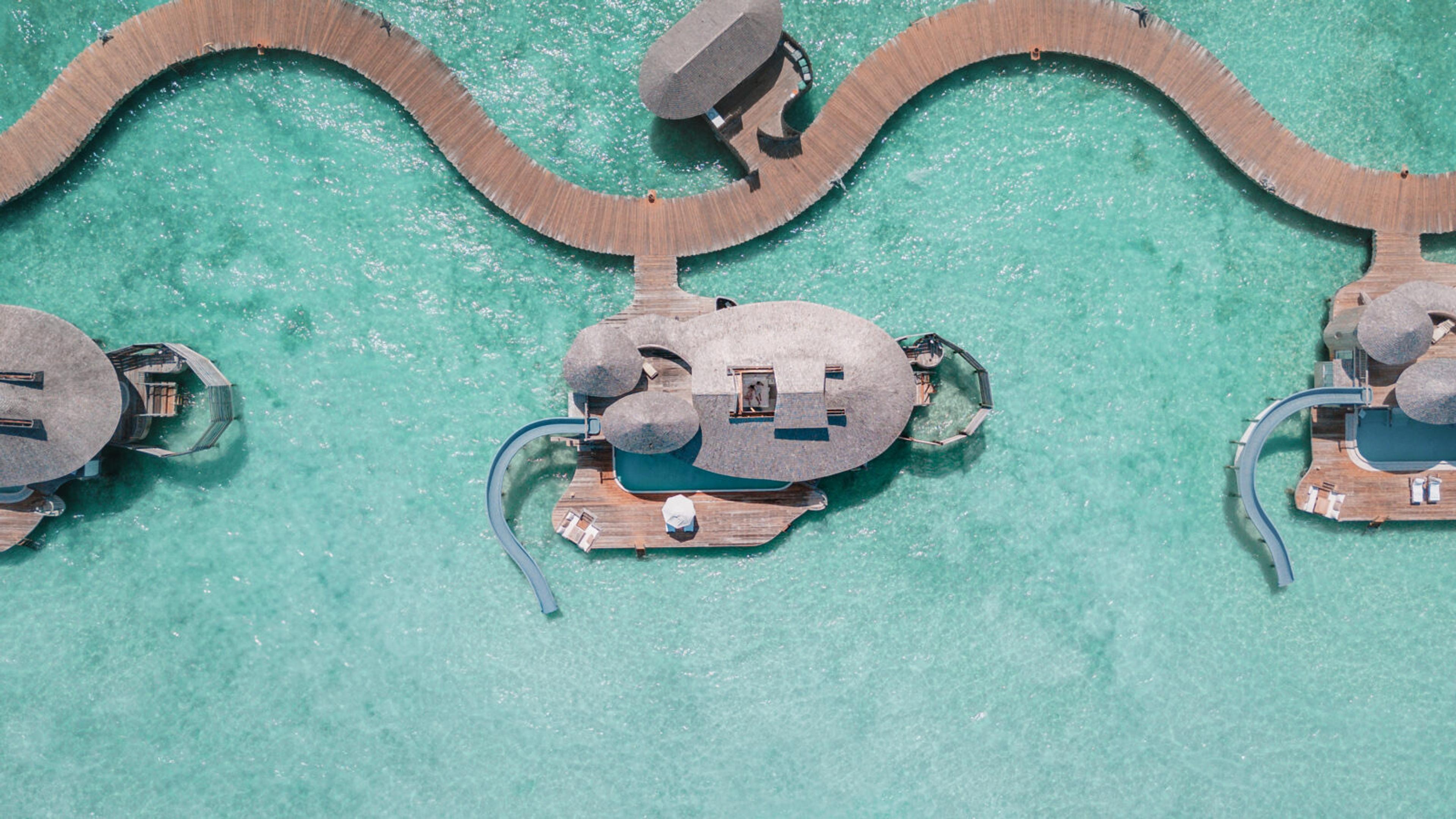 a cluster of overwater villas with thatched roofs, linked by a winding wooden walkway over turquoise lagoon water, seen from directly above