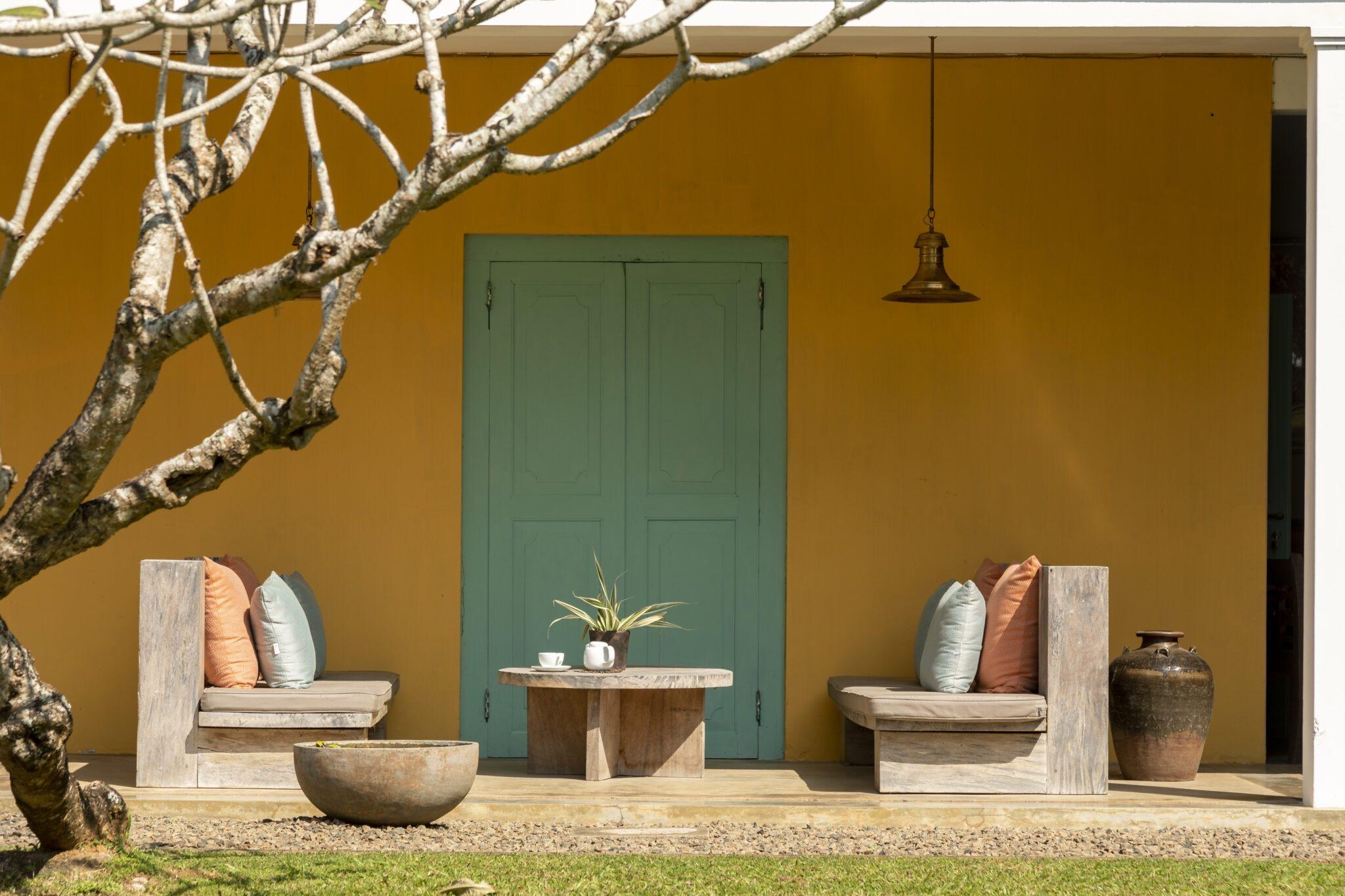 Two low wooden benches with pink and sage cushions face each other over a round stone table, in front of a mustard wall and a teal double door, a bare tree in the foreground.