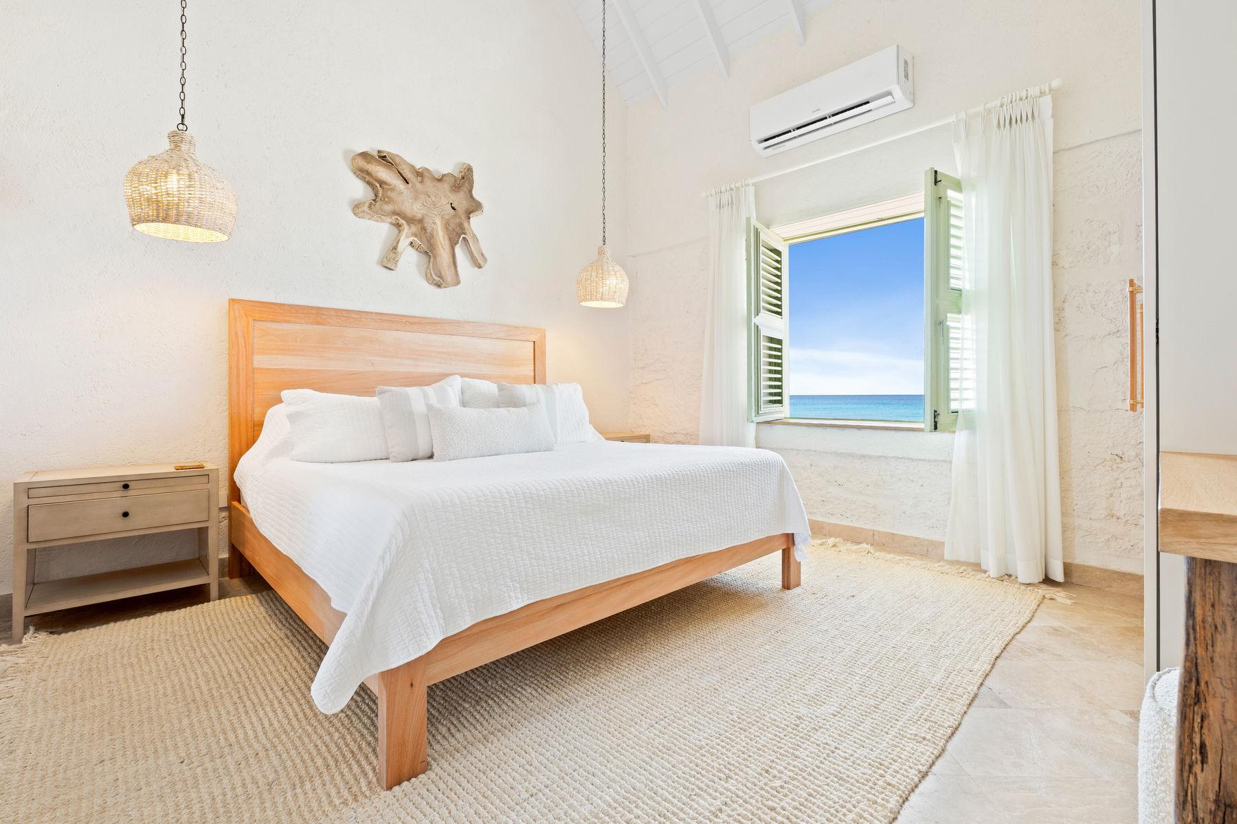 A wood framed bed with white linen beneath a driftwood wall hanging, a window open to the sea.