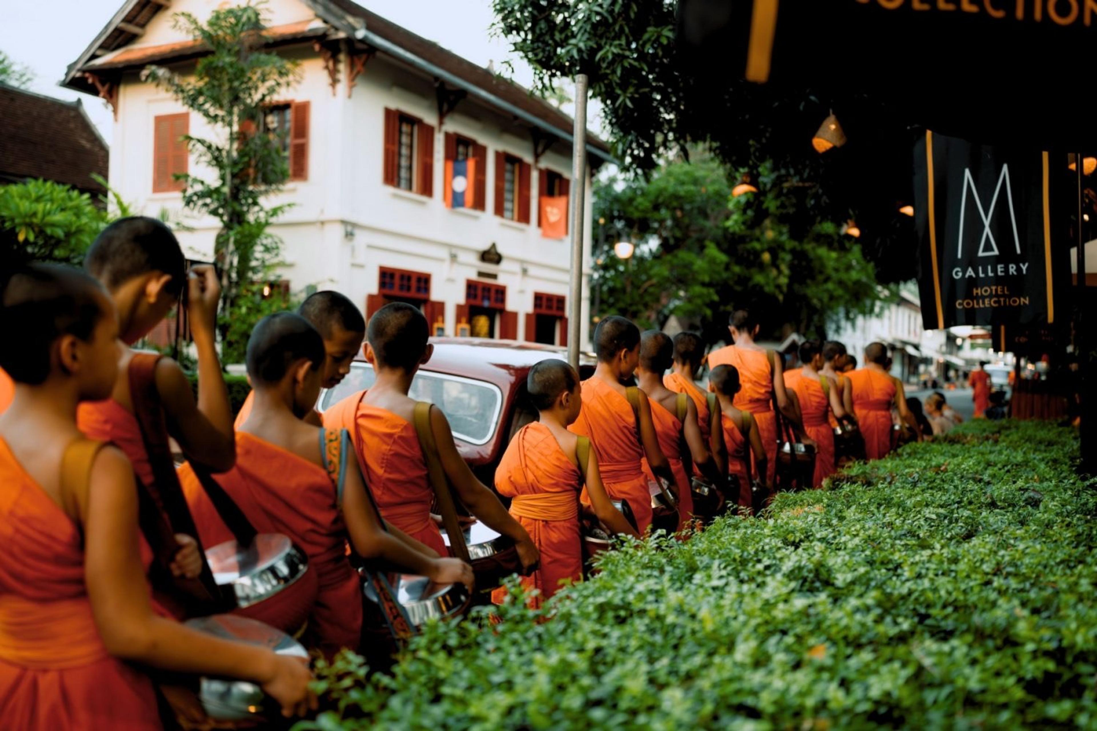 A line of young novice monks in orange robes walk along a street carrying alms bowls, past hedges with the hotel's white facade visible behind.