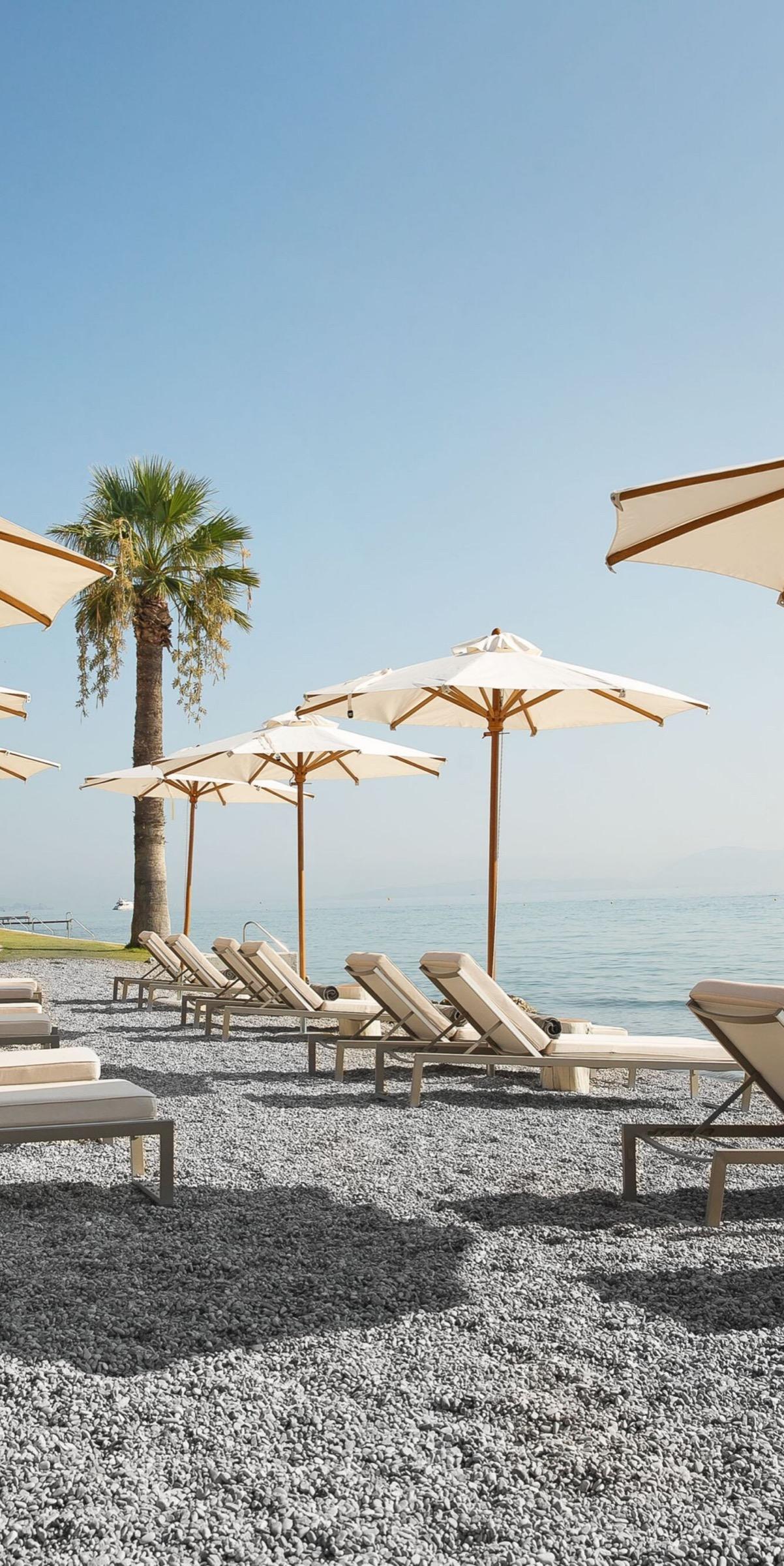 Empty sunloungers and cream umbrellas on a pebble beach beside the sea, with a palm tree and a line of further umbrellas along the shore.