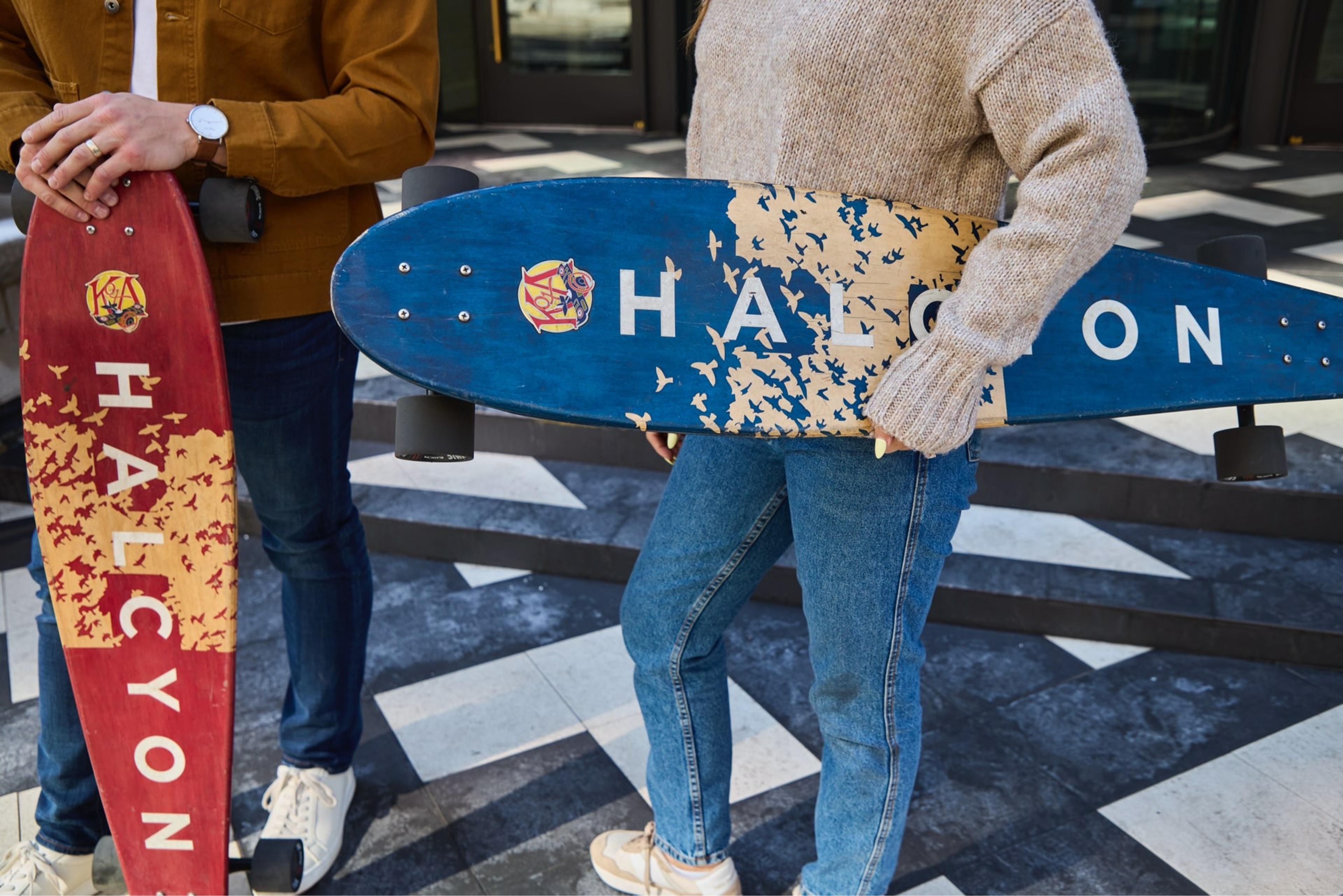 Two people, cropped above the face, hold longboards printed with the Halcyon name in front of a glass entrance, on a black and white tiled plaza.